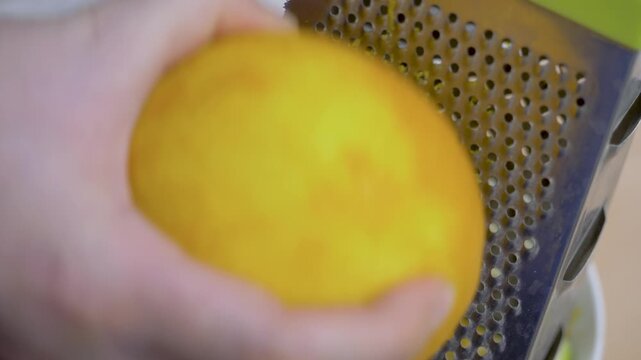 Female baker hand holds grater steady and rubs ripe orange into fine pieces. Peel turns to zest for cake mix. Precision, patience, and culinary discipline. Shredder kitchen utensil used to shred foods