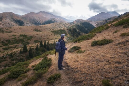 Hiker with backpack standing on a mountain trail overlooking Dzungarian Alatau range under cloudy sky in Kazakhstan. Commercial background for outdoor adventure, hiking tourism, and active lifestyle 