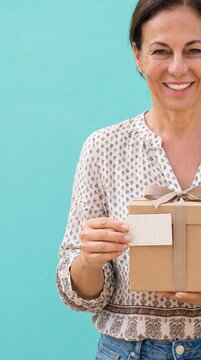 Smiling Middle Aged Woman Holding a Gift Box with a Blank Tag Against a Teal Background