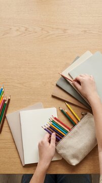 Hands organizing colored pencils and notebooks on a wooden desk for creative work or study