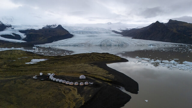Aerial view of bubble hotel pods situated on a black sand shore near a glacier lagoon with floating icebergs and snow-capped mountains in the background, Vik, Myrdalshreppur, Iceland.