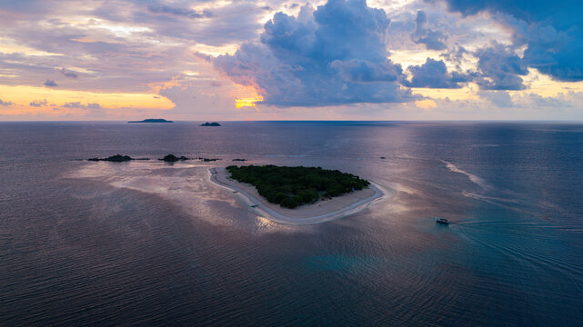Aerial view of a small tropical island with lush vegetation and a white sand beach surrounded by the deep blue sea under a dramatic sunset sky in Busuanga, Mimaropa, Philippines.