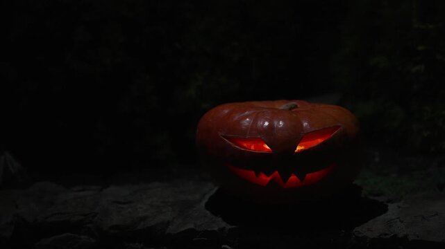 Menacing carved pumpkin face with glowing eyes on rough stone plinth in night darkness, super slow motion footage with negative copy space