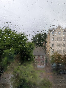 Abstract view of raindrops on a glass pane with an overcast, moody bokeh of green trees and urban buildings in the background.