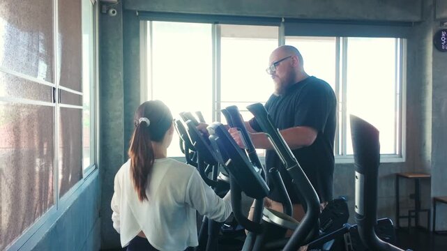 A fitness trainer coaches a woman on an elliptical machine in a bright gym, pointing at the console during a cardio workout.