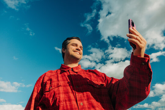 Smiling man in red plaid shirt taking selfie outdoors, smartphone, blue sky, low angle
