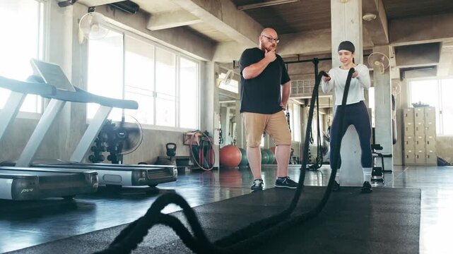 Woman works battle ropes in a bright gym while a trainer guides her near treadmills and equipment.