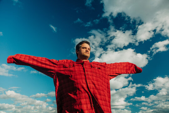 Man in red plaid shirt with open arms, confidence, optimism, dramatic cloudy sky, low angle