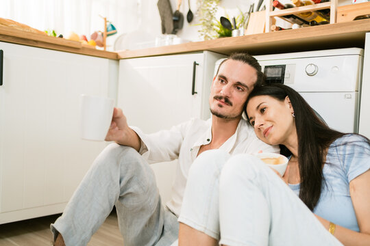 Couple sitting on a kitchen floor with mugs, leaning together and relaxing by the dishwasher, enjoying a calm coffee break in a bright home