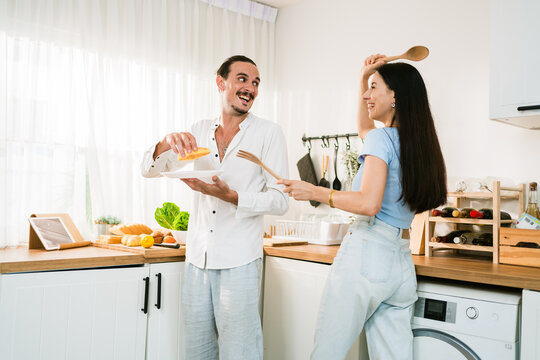 A playful couple cooks in a bright modern kitchen, smiling and waving wooden utensils while preparing food by the counter near a window and washer.