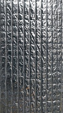 ​A vertical close-up shot of a silver foil thermal insulation sheet featuring a embossed grid pattern with shiny light highlights and dark metallic reflections
