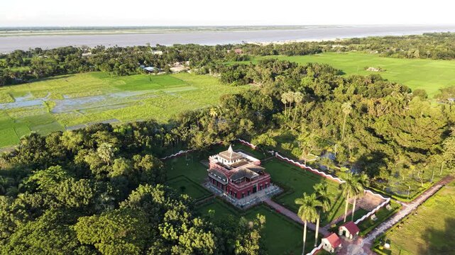 A circular drone orbit reveals different perspectives of Shilaidaha Rabindra Kuthibari, highlighting its long red facade, rows of columns, leafy surroundings, and the distant river landscape.