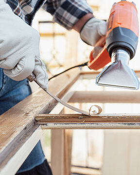 Adult carpenter wearing protective leather gloves, using a heat gun and a wood scraper to remove paint from an old wooden window. Construction industry, carpentry. Restoration. 