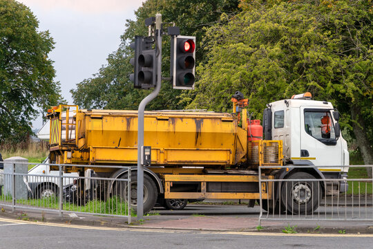 A yellow commercial road-repair, maintenance truck  stopped on a paved surface next to a roadside pedestrian barrier, positioned underneath a set of traffic lights. Is fitted with an asphalt hotbox.