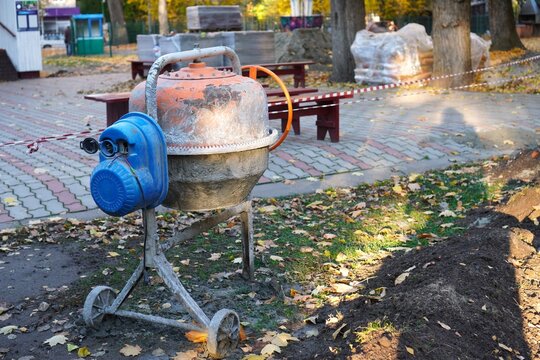 Cement mixer on autumn park renovation site near paved walkway and bench. Excavated soil, warning tape and fallen leaves, seasonal city landscaping and pathway repair works in October.