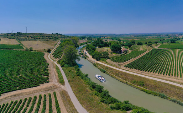 Aerial view of Canal du Midi with a pleasure boat cruising through lush vineyards and rural landscape under a clear blue sky in Beziers, Occitanie, France.
