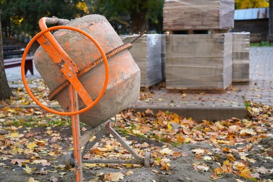 Orange cement mixer and stacked paving slabs on autumn park walkway renovation site. Seasonal sidewalk repair with organized safe work zone among fallen leaves, October landscaping works.