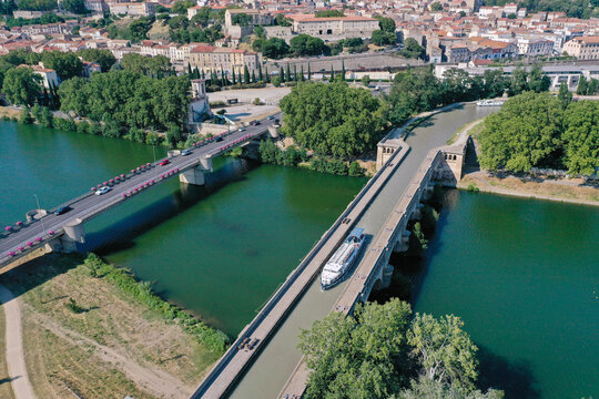 Aerial view of the Canal du Midi waterbridge crossing the Orb River with a boat navigating the canal, adjacent to a road bridge and the historic city of Beziers, Occitanie, France.