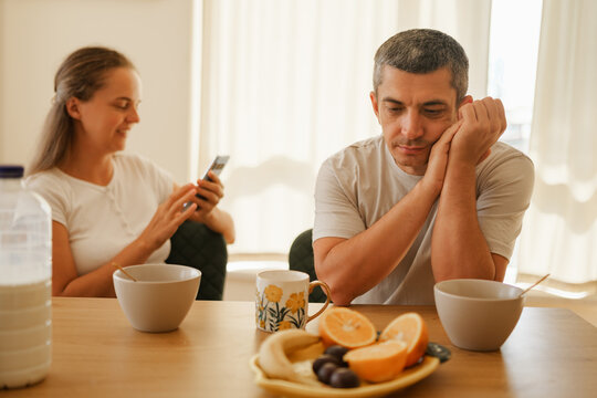 Bored husband sitting thoughtful at breakfast table while his wife ignores him using her smartphone in bright kitchen.