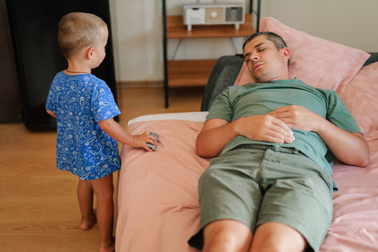 Little boy standing near a bed and playing with a toy car while his tired father sleeps on the bed at home.