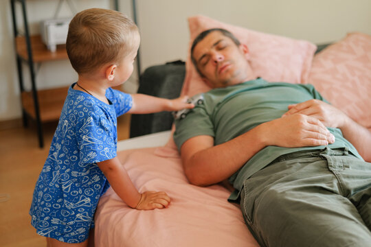 Little boy standing by the bed and playing with a toy car on his sleeping father's shoulder in a cozy bedroom.