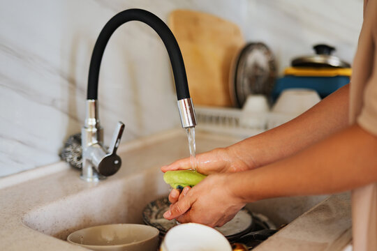Close-up of human hands washing dishes with a sponge under running water from a tap in a kitchen sink.