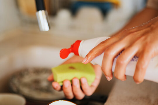 Close-up of human hands squeezing dishwashing liquid soap from a bottle onto a sponge in the kitchen sink.