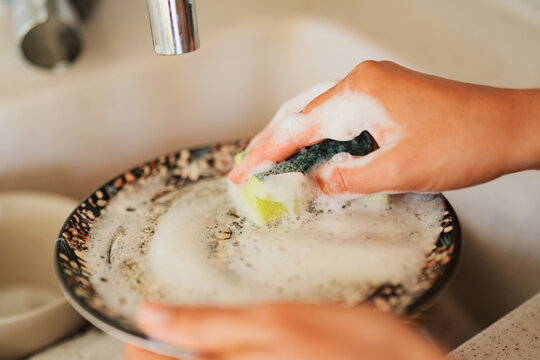 Close-up of human hands cleaning a ceramic plate with a soapy, foamy sponge over a kitchen sink.