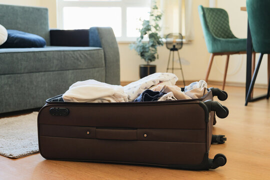 Open brown suitcase packed with clothes lying on a wooden floor in a modern living room.