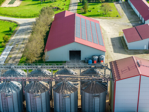 Aerial view of an agricultural facility featuring metal grain silos and a large warehouse with solar panels on its red roof in Lode, Sigulda Municipality, Latvia.