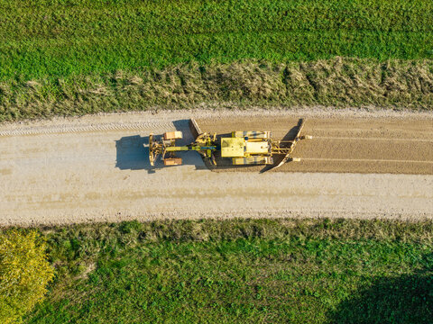 Aerial view of a yellow motor grader leveling and reshaping the surface of a rural gravel road in Lode, Sigulda Municipality, Latvia.