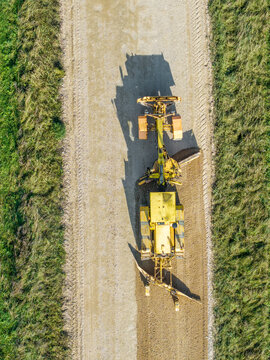 Aerial view of a yellow motor grader leveling and reshaping the surface of a rural gravel road with its blade in Lode, Sigulda Municipality, Latvia.