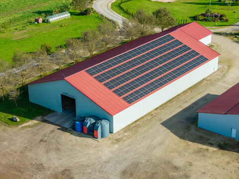 Aerial view of a warehouse with solar panels installed on its red roof, surrounded by green fields and gravel grounds in Lode, Sigulda Municipality, Latvia.