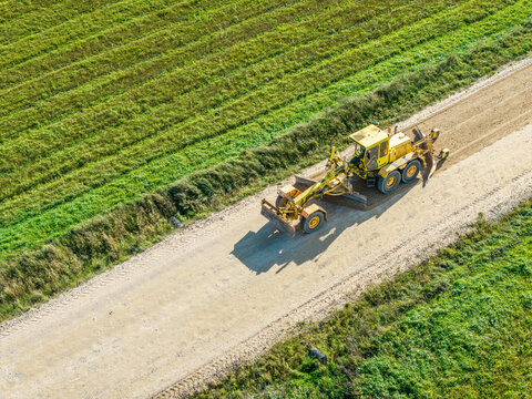 Aerial view of a yellow motor grader leveling and reshaping the surface of a rural gravel road surrounded by green fields in Lode, Sigulda Municipality, Latvia.