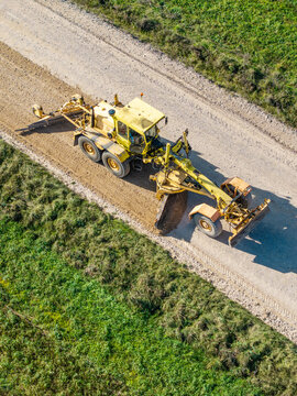 Aerial view of a yellow motor grader leveling and reshaping the surface of a rural gravel road in Lode, Sigulda Municipality, Latvia.