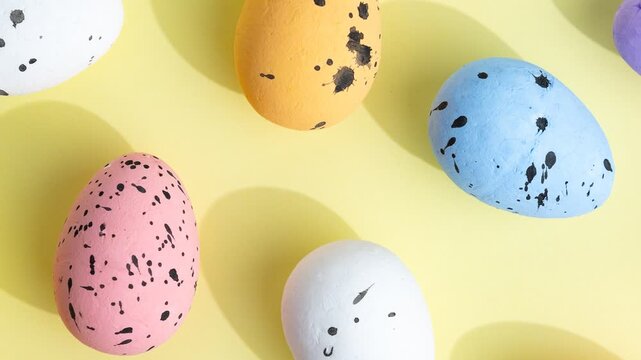 Pastel-colored Easter eggs with black speckles arranged on a soft yellow background. Vibrant shades of purple, blue, white, orange, and pink create a festive and cheerful scene perfect for Easter.