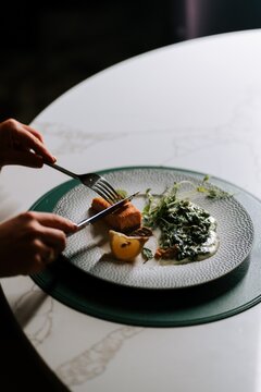 Person cutting a piece of grilled salmon with spinach and lemon on a white marble table for dinner
