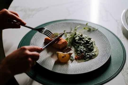 Person cutting a piece of cooked salmon served with creamy greens and a fresh lemon slice on a plate