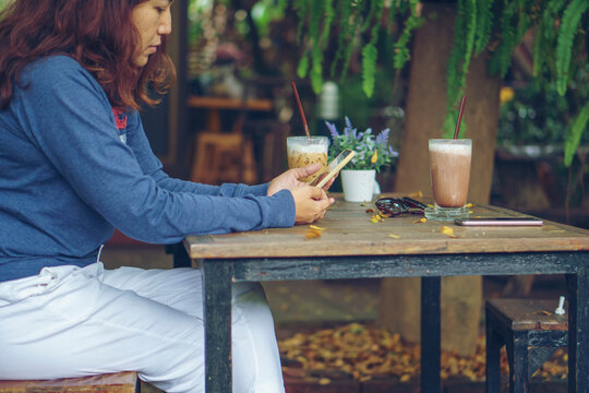 Lifestyle asian woman using smartphone at wooden cafe table with iced coffee and chocolate drink, sunglasses, and potted flowers, enjoying quiet leisure time in outdoor garden.
