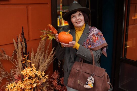Stylish woman in hat holding orange pumpkin and maple leaves near house entrance decorated with dried flowers and pampas grass. Natural eco autumn decor for Thanksgiving and Halloween holidays.