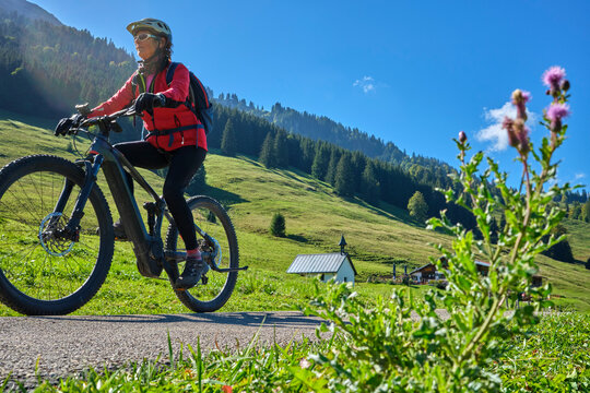 Active senior woman cycling on an electric mountain bike across an alpine meadow with forested slopes and mountain peaks under a blue sky . Allgaeu Alps, Steibis-Oberstaufen, Bavaria, Germany