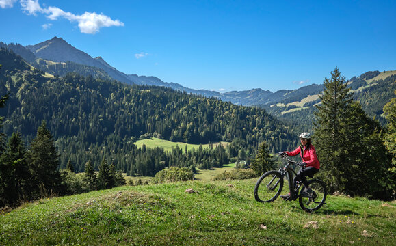 Active senior woman cycling on an electric mountain bike across an alpine meadow with forested slopes and mountain peaks under a blue sky . Allgaeu Alps, Steibis-Oberstaufen, Bavaria, Germany