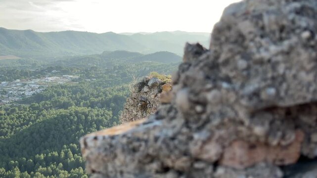 Wide angle static shot looking past ancient stone ramparts toward the lush green valley and rolling mountains in Xativa, Valencia, Spain.