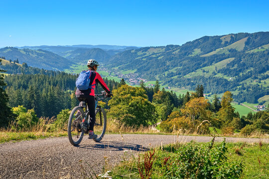 Female mountain biker overlooking a scenic alpine valley with green hills, dense forests and distant villages on a sunny day. Thalkirchdorf, Bavaria, Germany