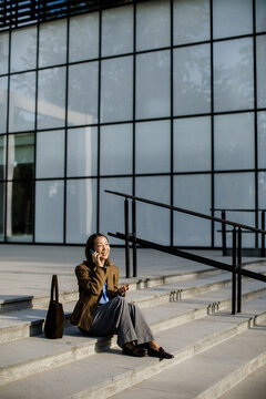 Woman sitting on steps talking on phone near modern building