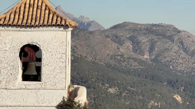 Smooth panning shot of the historic white bell tower and tiled roof overlooking the majestic mountain landscape under soft winter sunlight in Montanejos, Spain.