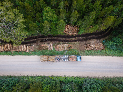 Aerial view of a logging truck with a hydraulic crane loading cut timber logs onto a trailer along a forest road in Latvia.