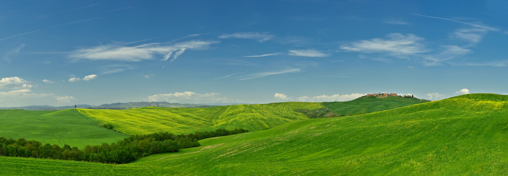 Aerial view of rolling green hills and a distant hilltop village under a bright blue sky in Tuscany, Italy.
