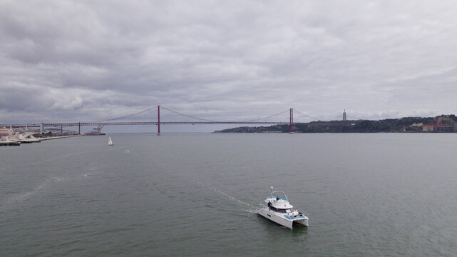 Aerial view of a white catamaran sailing on the Tagus River with the 25 de Abril Bridge in the background under a cloudy sky in Lisbon, Lisbon, Portugal.