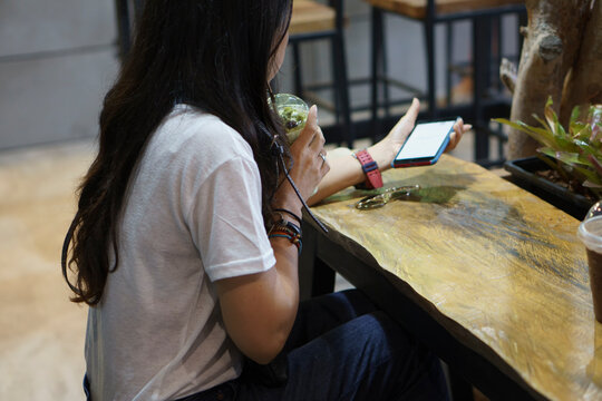 Woman side view using smartphone and holding iced coffee at cafe. Female sitting and enjoy relaxing time indoor. Lifestyle and technology concept.